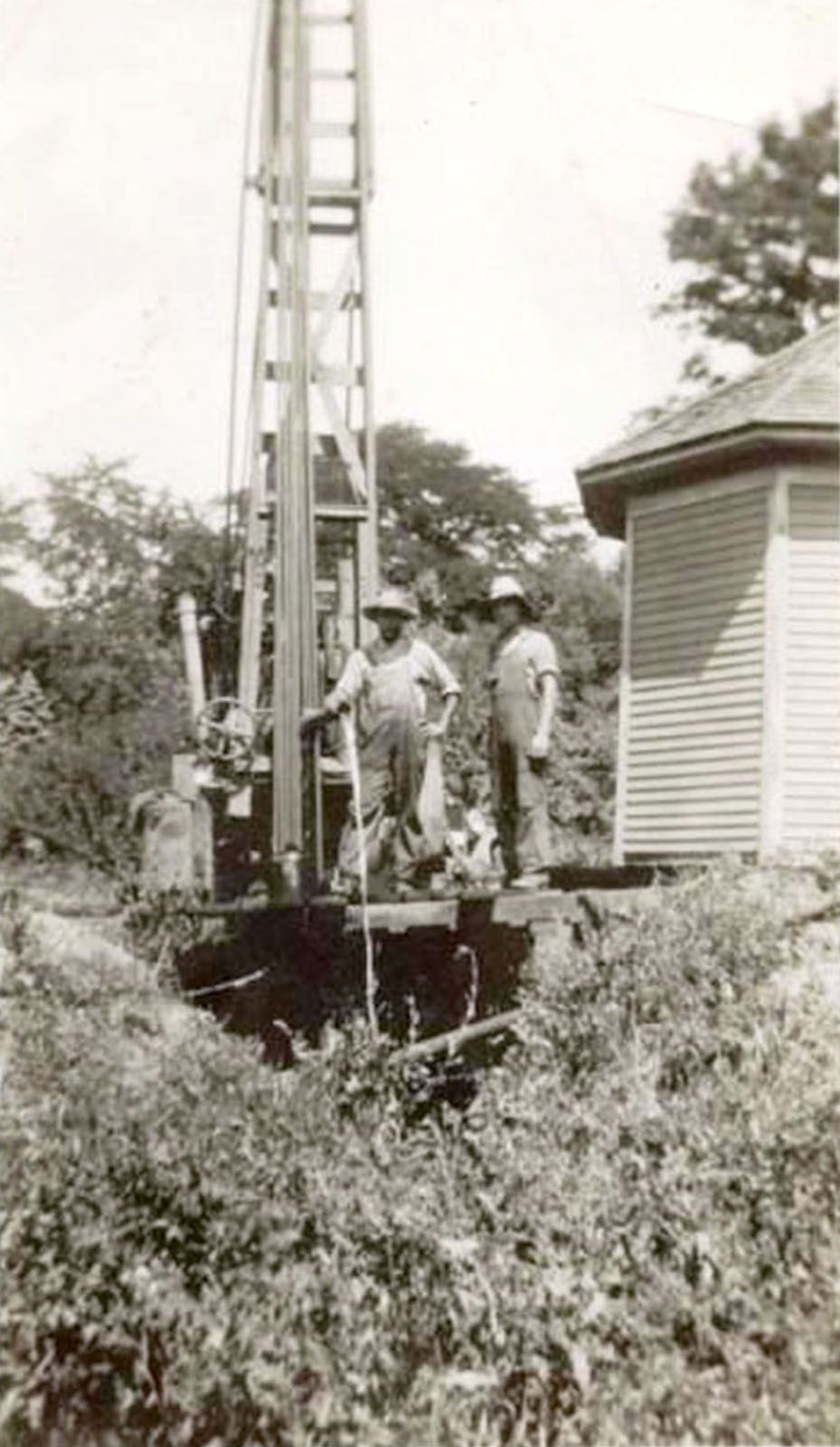 Workers beside a tall drilling rig near a small house