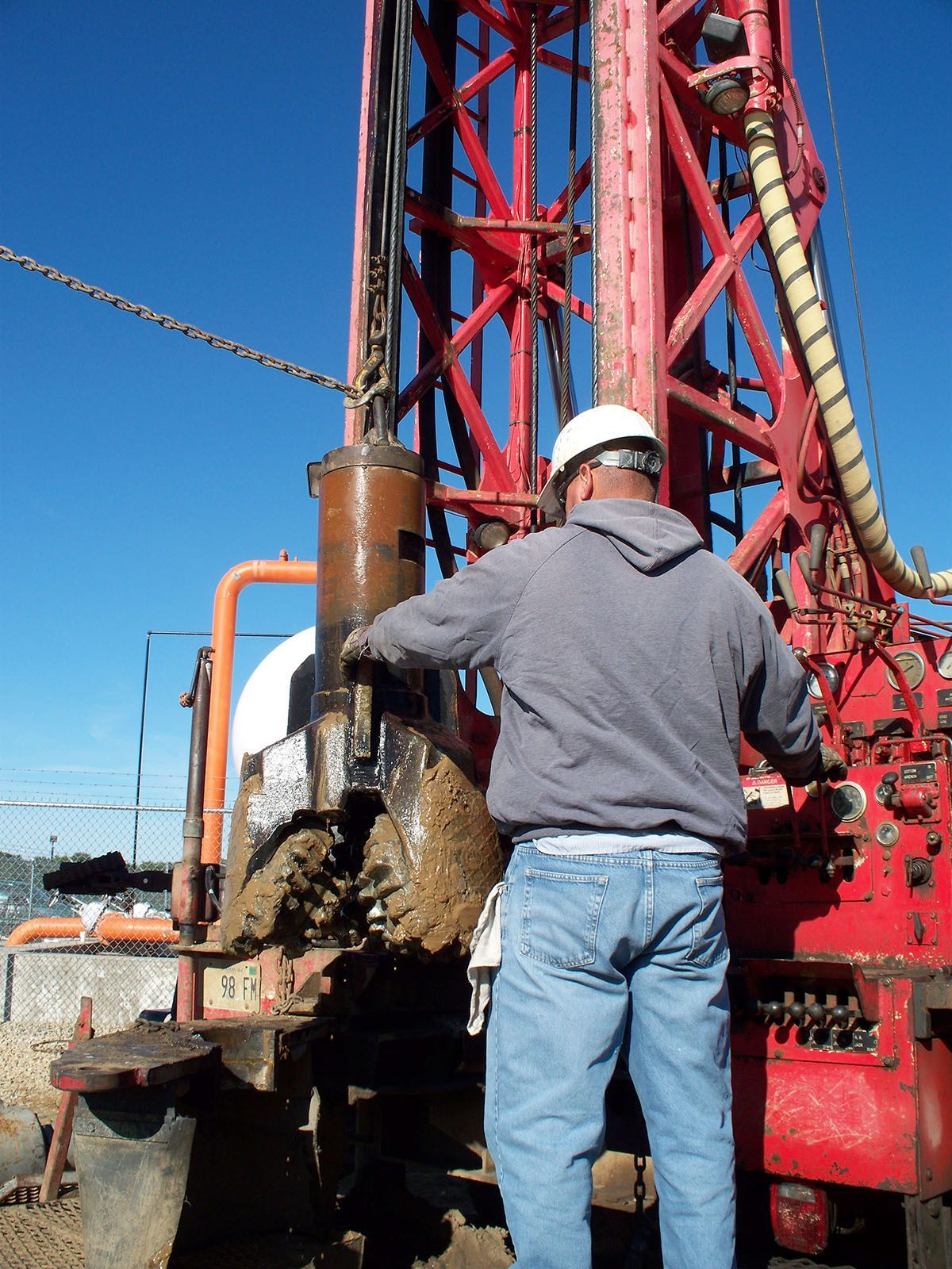 Worker in a white hard hat stands beside a red drilling rig