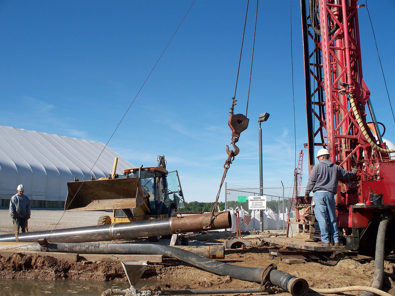 Construction site with drilling rig, workers, and heavy equipment beside a large pipe