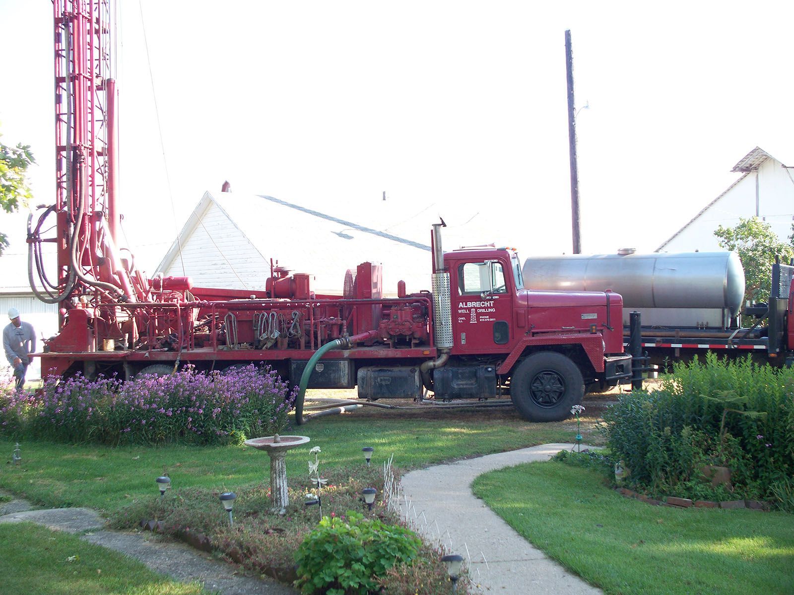 Red drilling truck beside a house, with a tall rig and a silver tank in a grassy yard