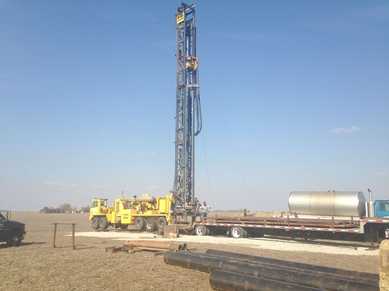 Yellow drilling rig and truck in a flat, sandy construction site under a clear blue sky