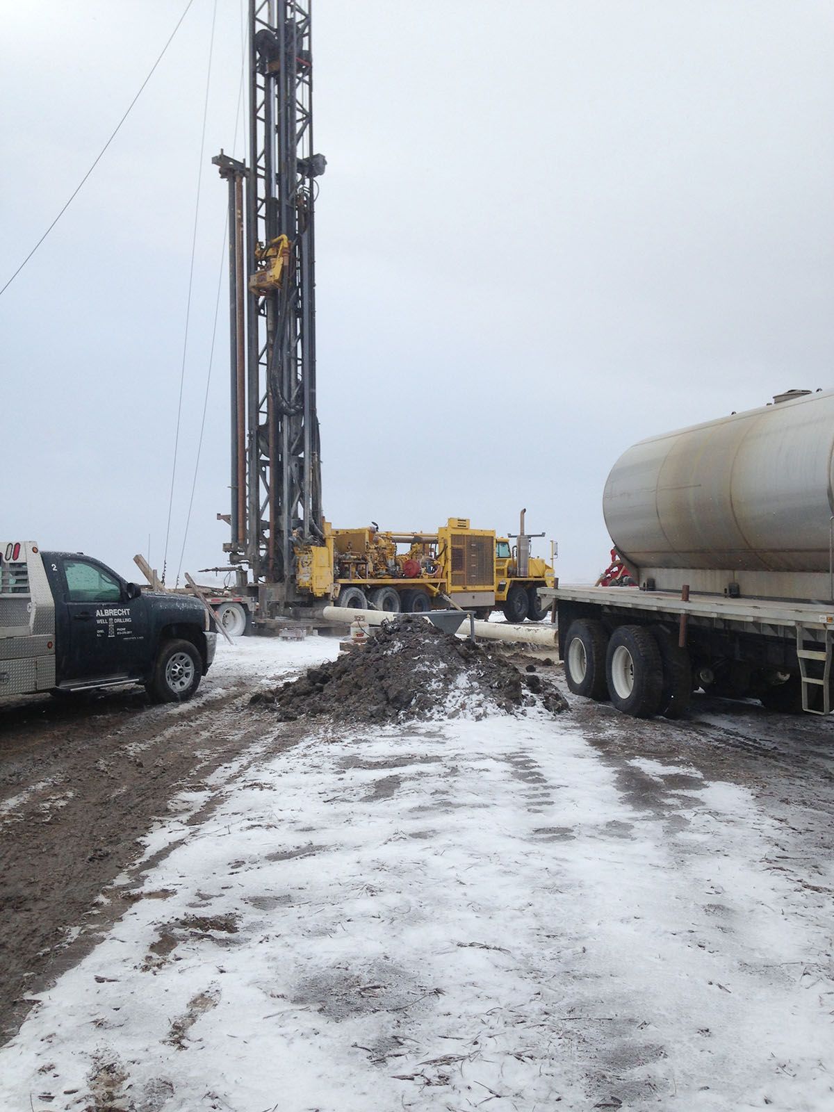 Snowy drilling site with a tall rig, trucks, and heavy equipment