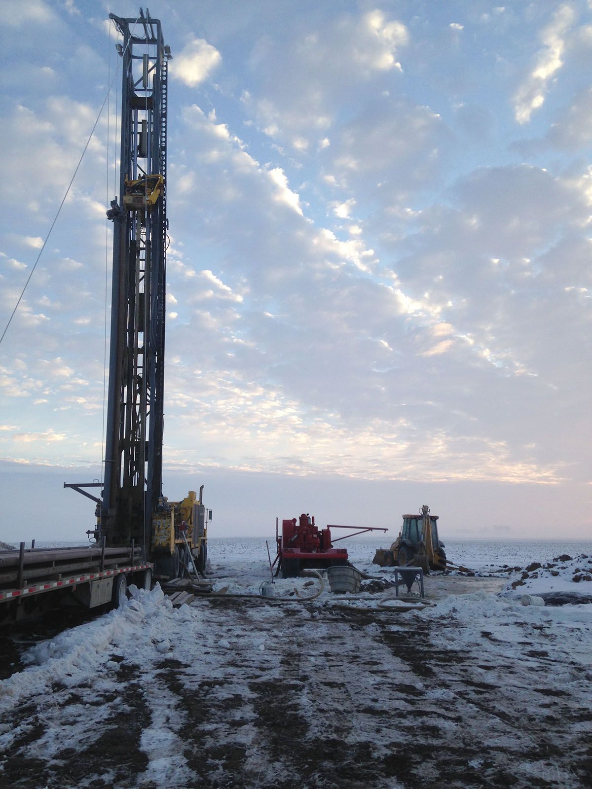 Snowy industrial site with tall drilling rig and machinery at dusk under a cloudy sky
