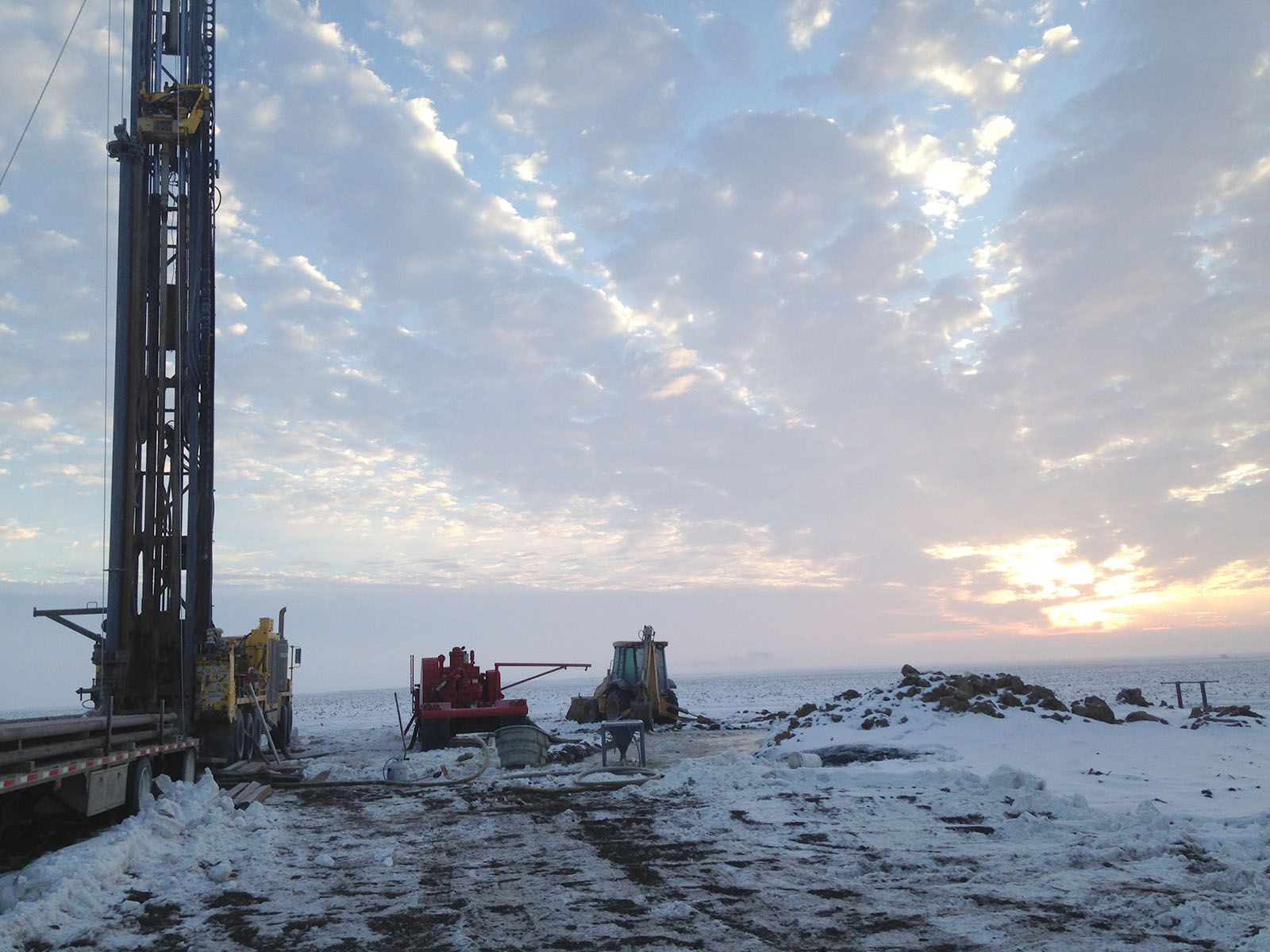 Snow-covered drilling site at sunset with tall rig and two work vehicles