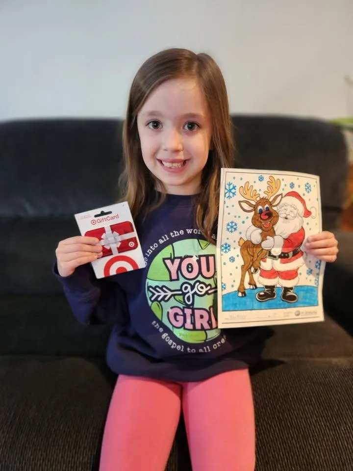 A smiling child holds a Target gift card in one hand and a colored picture of Santa and a reindeer in the other.