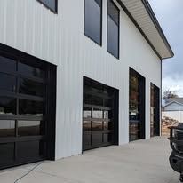 White metal building exterior with three black garage doors and a concrete driveway under construction.