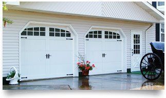 Two white garage doors on a house with a potted flower and a bicycle wheel at right