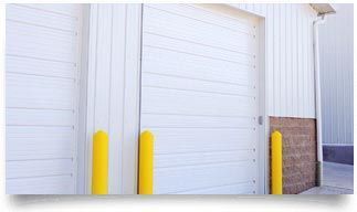 White warehouse loading dock doors with yellow safety bollards and a brick corner wall