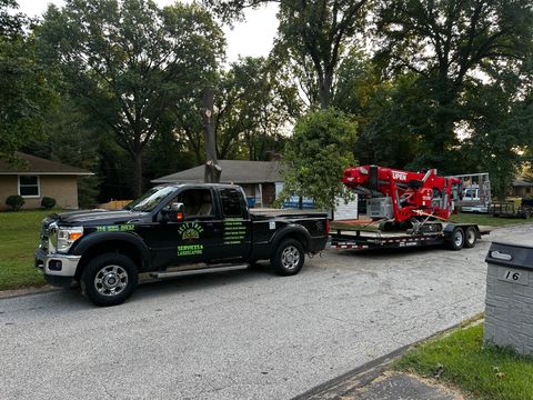 Black tow truck hauling a red boom lift on a trailer along a residential street