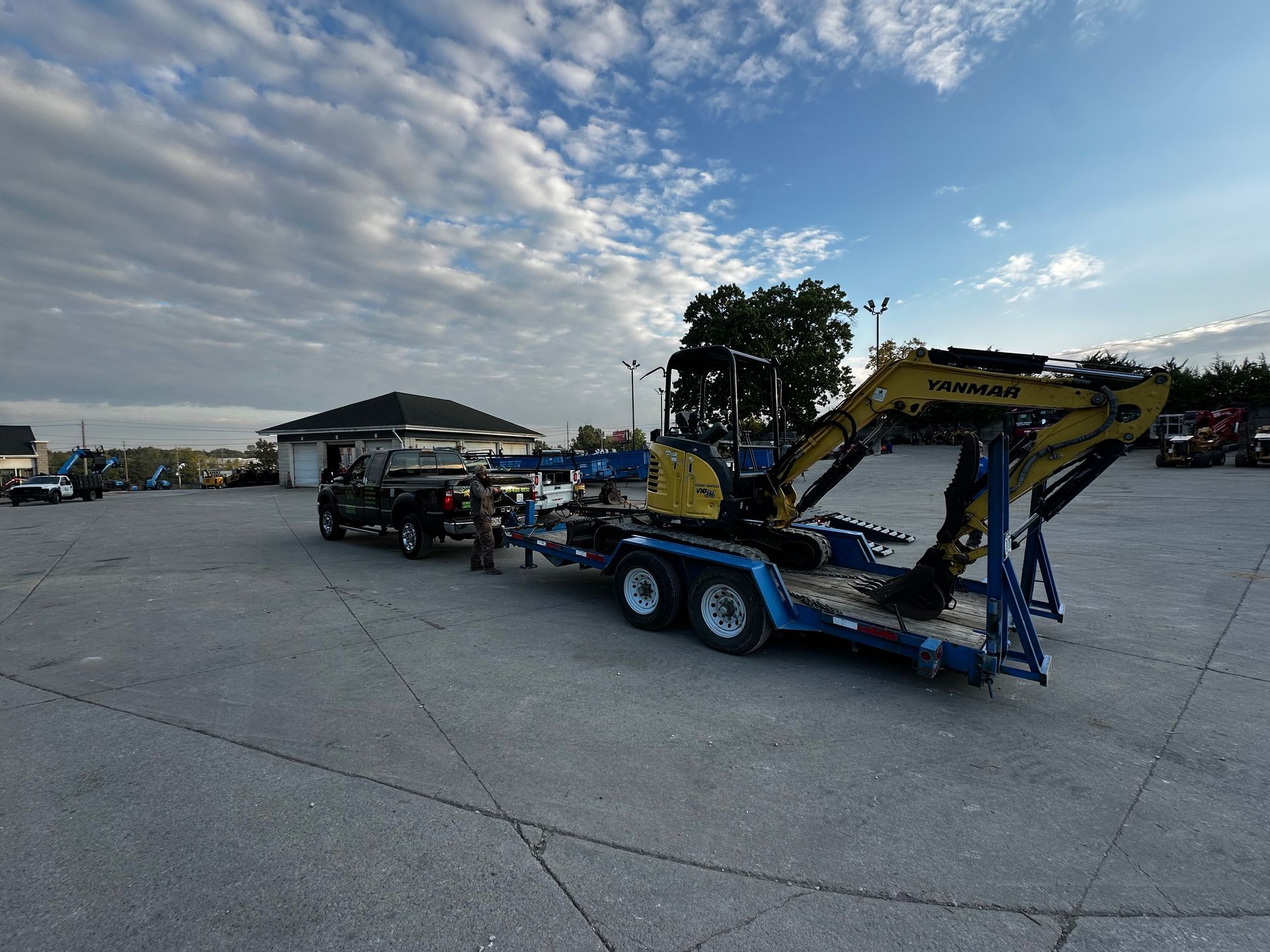 Blue tow truck carrying a yellow excavator in a large open parking lot under a cloudy sky
