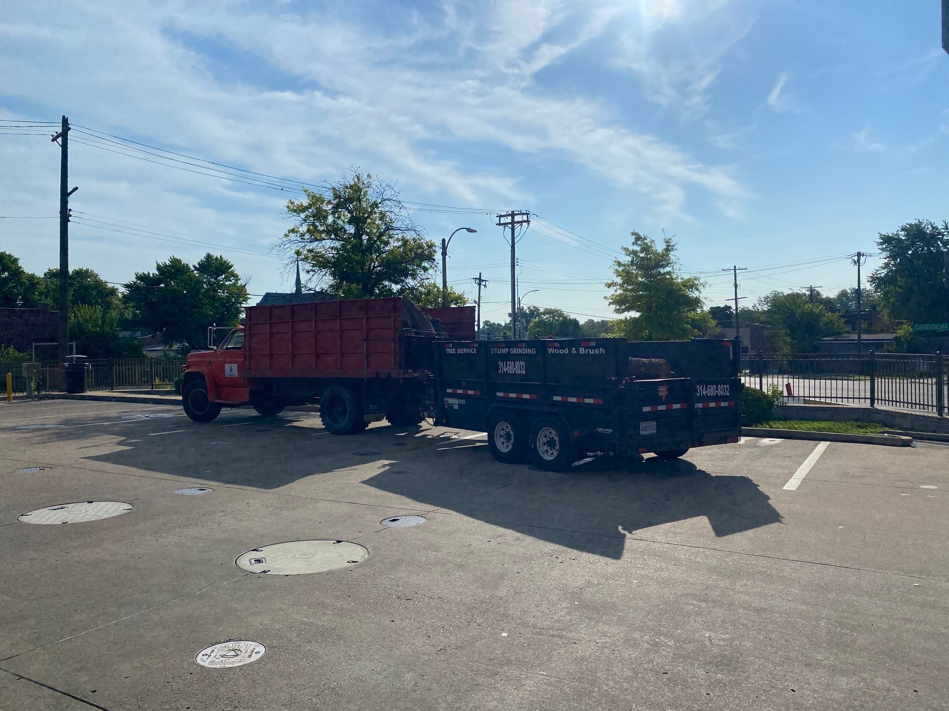 Two parked dump trucks in a sunny lot, one red and one black, near a fence and trees