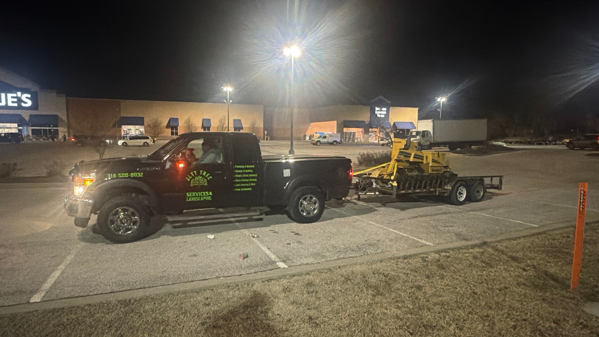 Black pickup truck towing yellow equipment trailer in a lit parking lot at night