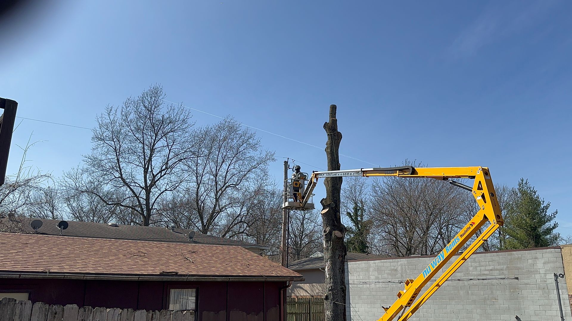 Yellow lift arm reaching above a wall beside a rooftop and bare trees under a blue sky