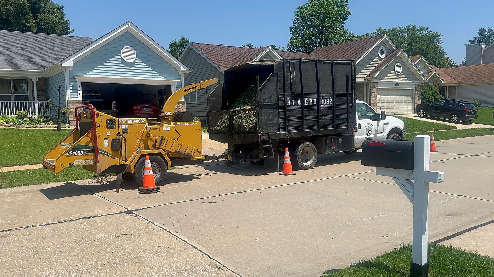 Yellow skid steer and black dump trailer in a suburban driveway with cones, workers, and an open garage.