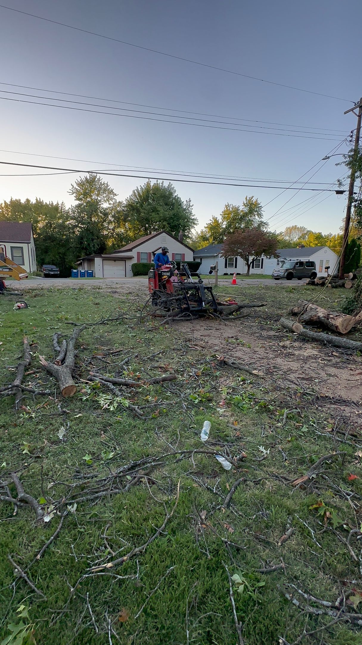 A person using a lawn tractor clears branches and debris in a yard at dusk, with houses in the background.