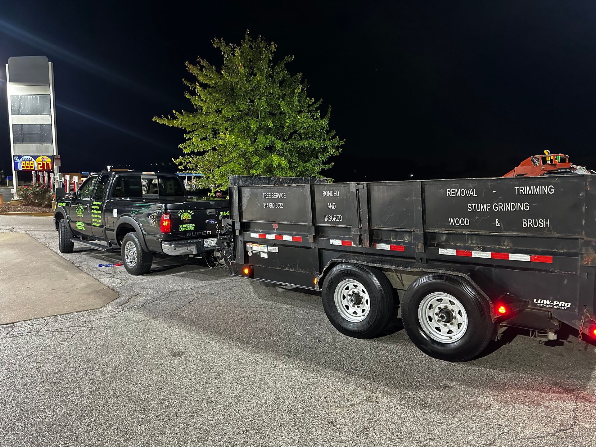 Black pickup truck towing a black dump trailer parked at night beside a lit building.