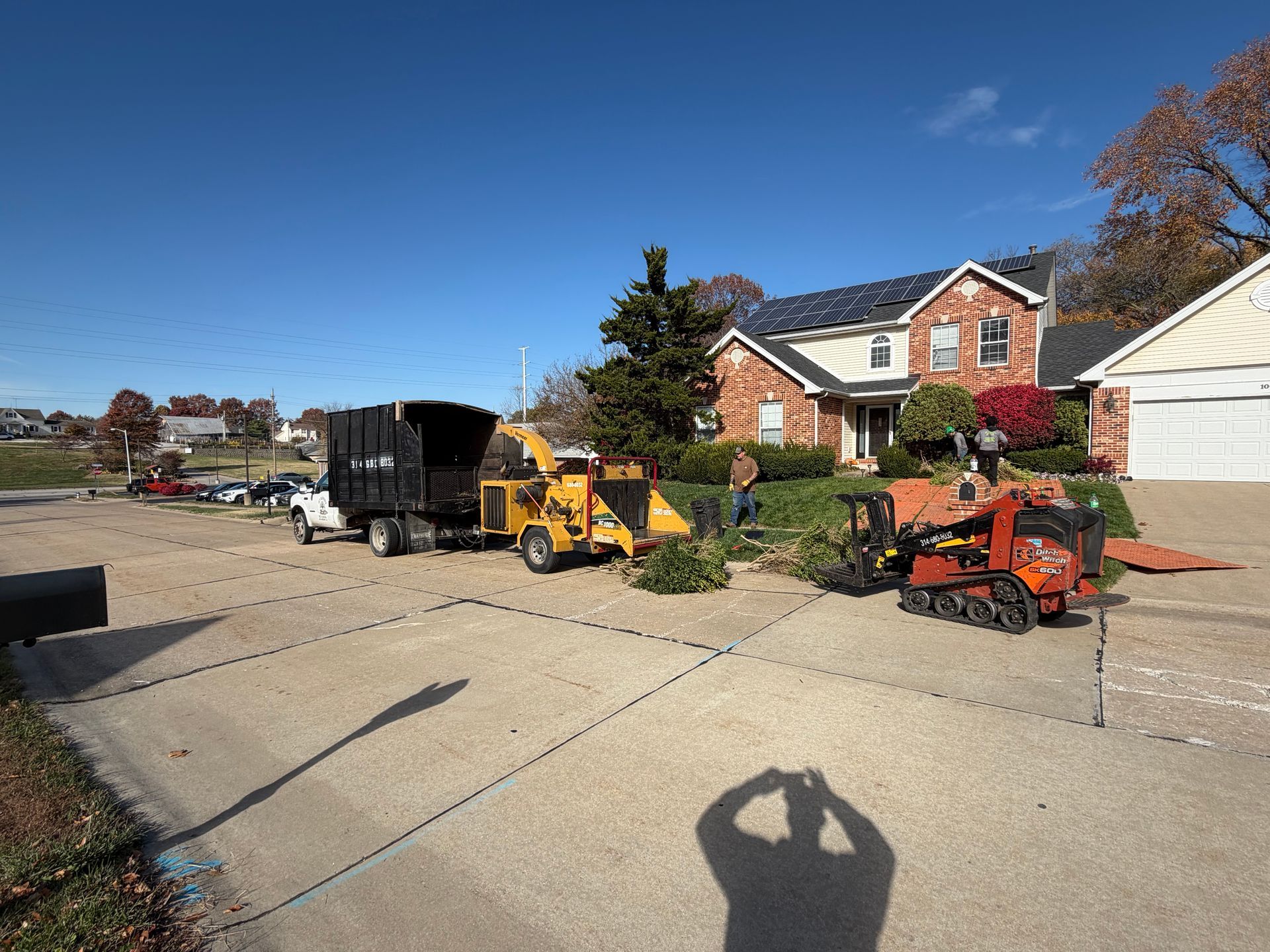 Construction equipment on a suburban street beside a brick house under a clear blue sky