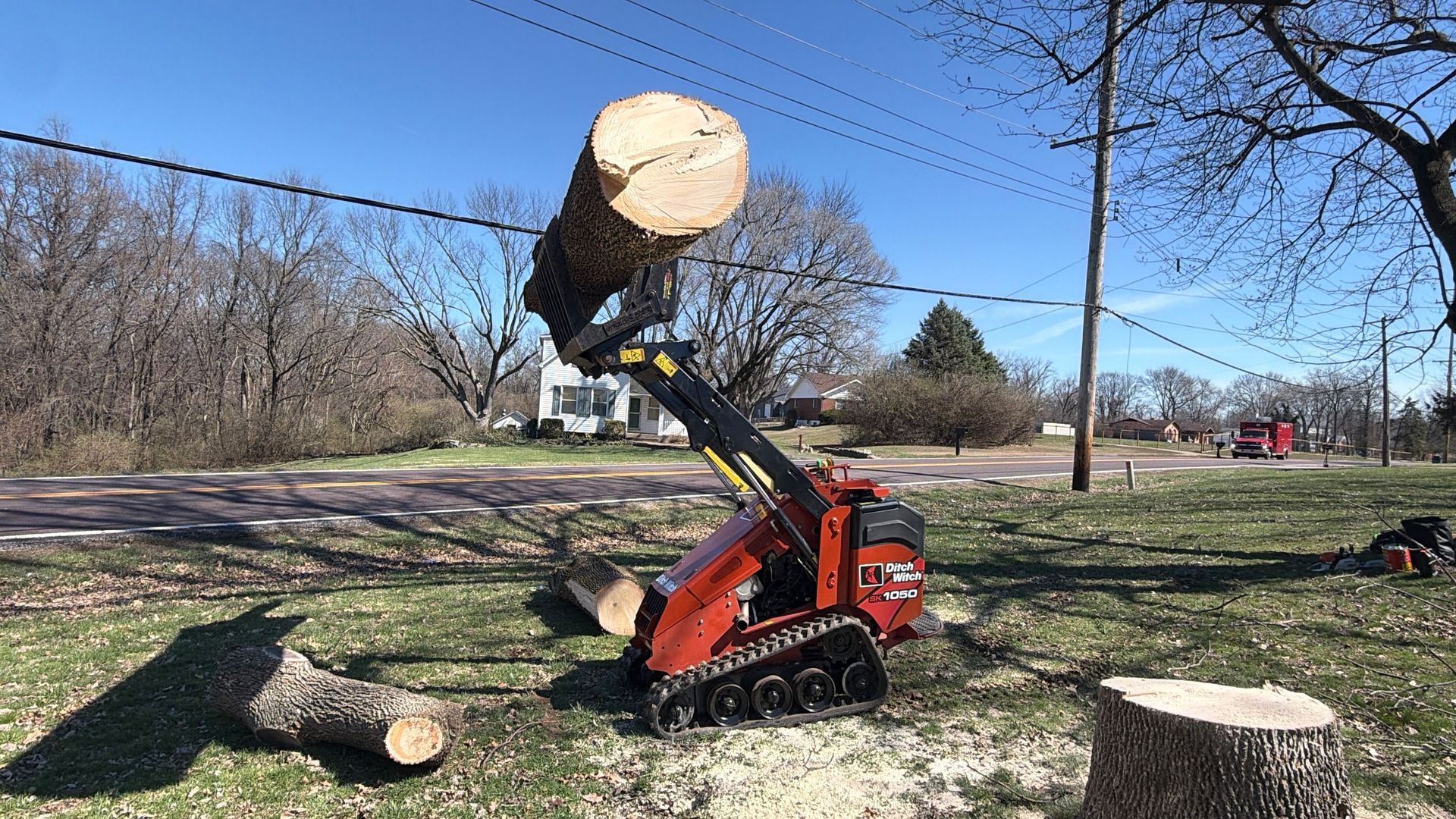 Excavator lifting a large cut tree trunk in a grassy yard with several log stumps and bare trees.
