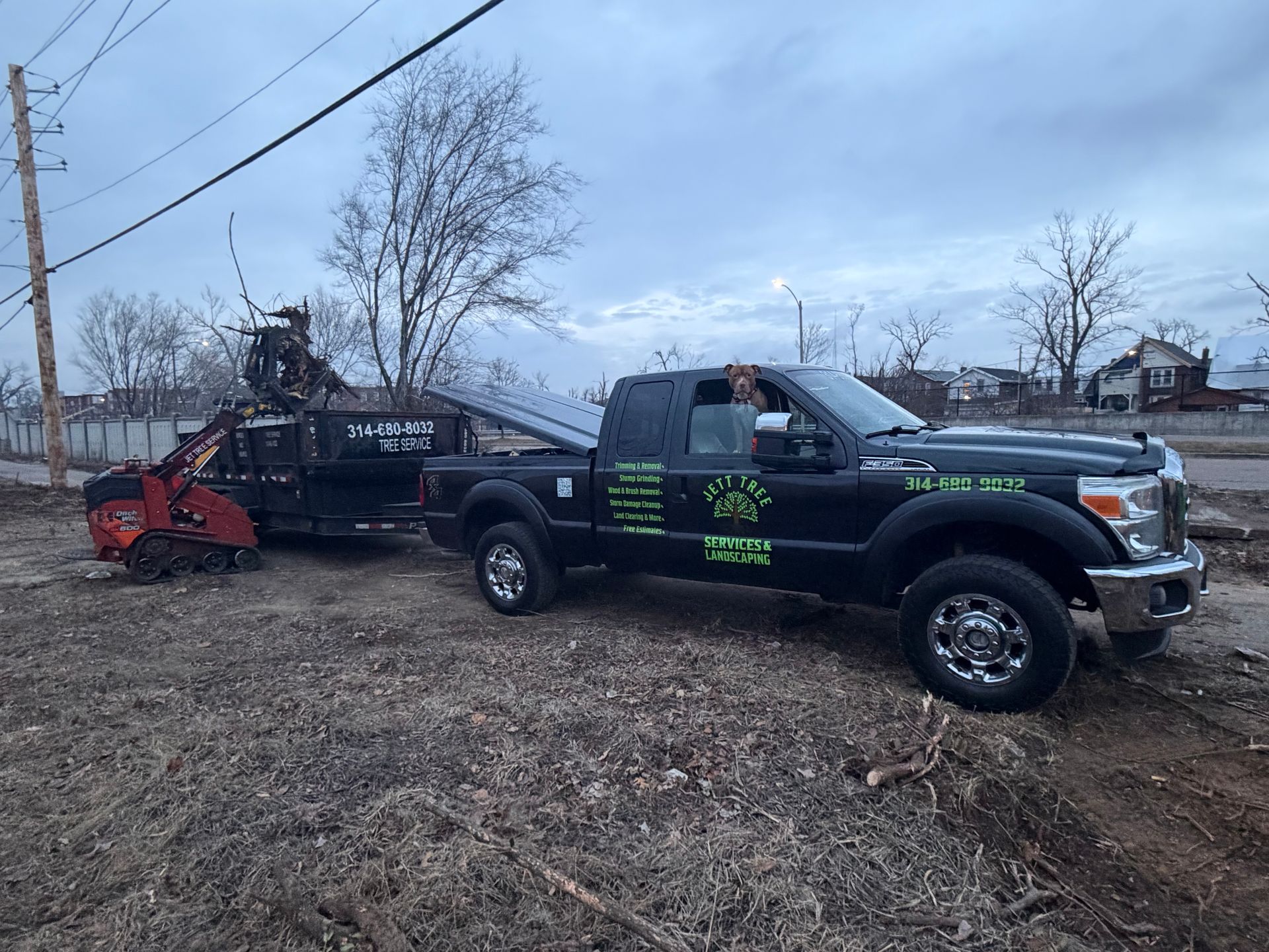 Black pickup truck hauling a trailer with a red skid steer on a muddy roadside under cloudy sky.