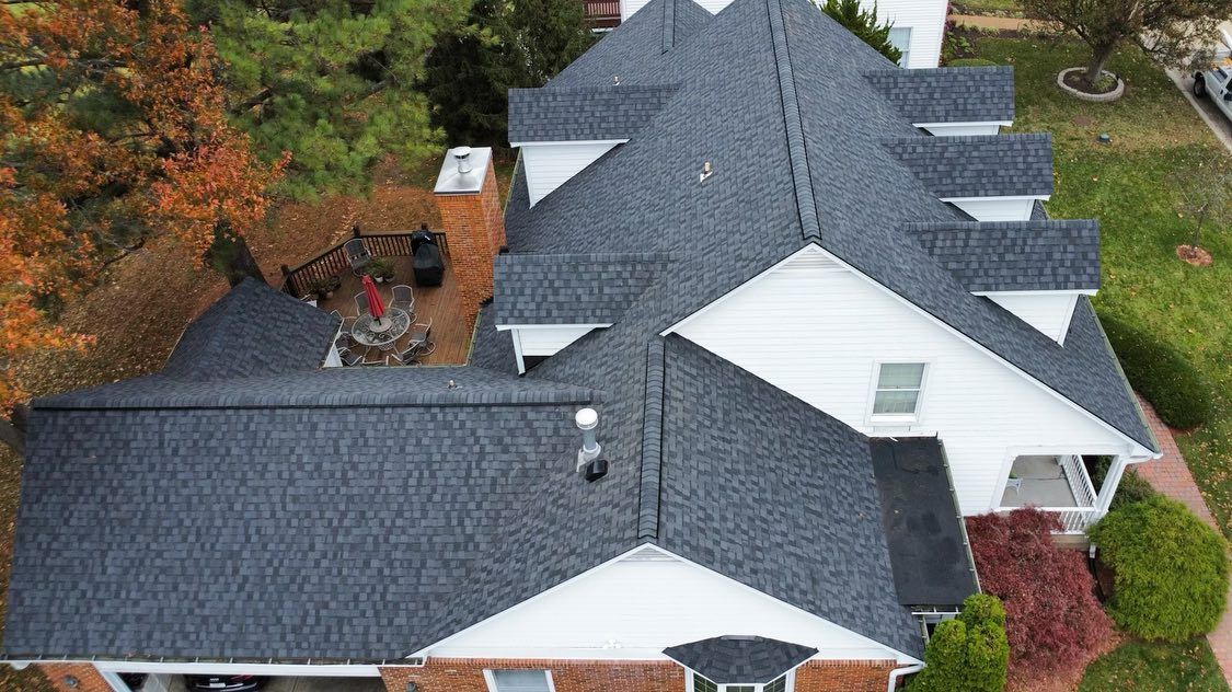 Aerial view of a dark gray shingled roof on a white house with dormers, set amid fall foliage.
