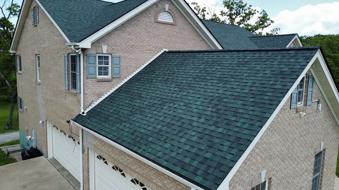 Beige brick house with a dark green shingled roof and white garage doors. Blue shutters are on a window.