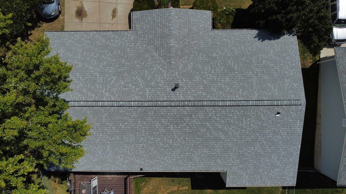Overhead view of a gray shingled roof with a ladder, situated in a residential area surrounded by trees and a driveway.