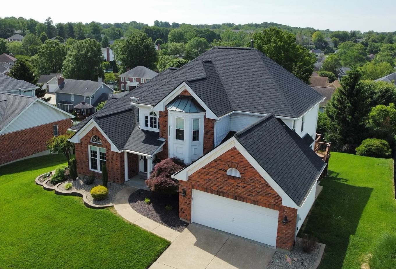 Aerial view of a two-story home with a black roof and red brick accents, set in a green, suburban neighborhood.