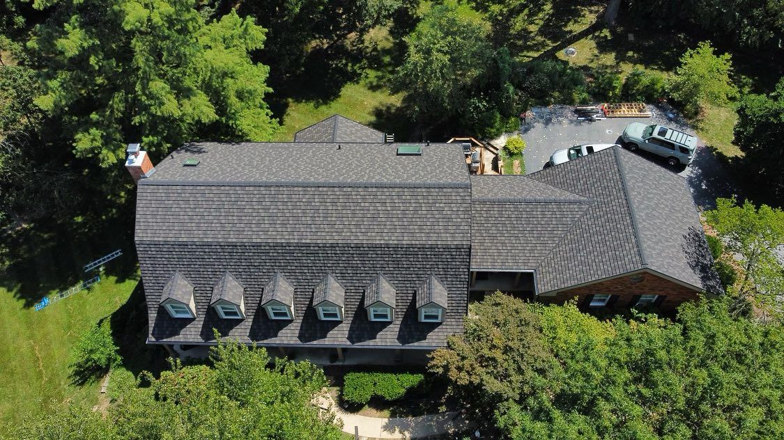Aerial view of a house with a dark gray roof, six dormer windows, and surrounding green trees.