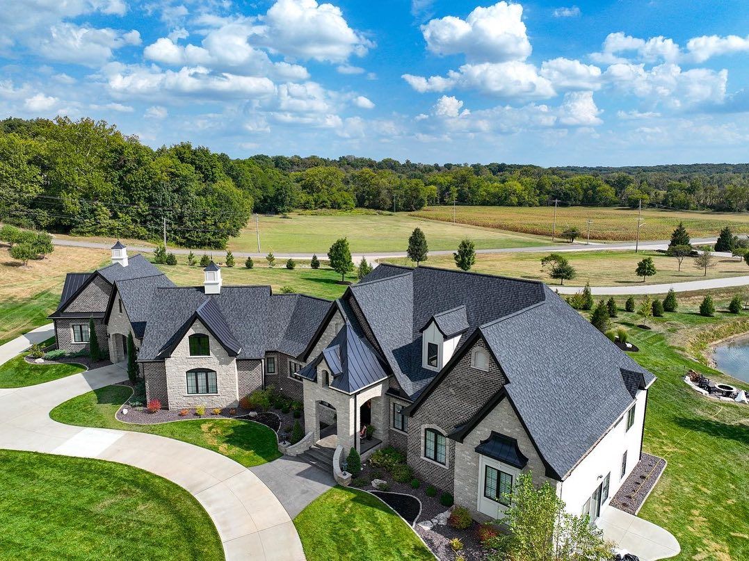 Large multi-roofed house with curved driveway and landscape under blue sky.
