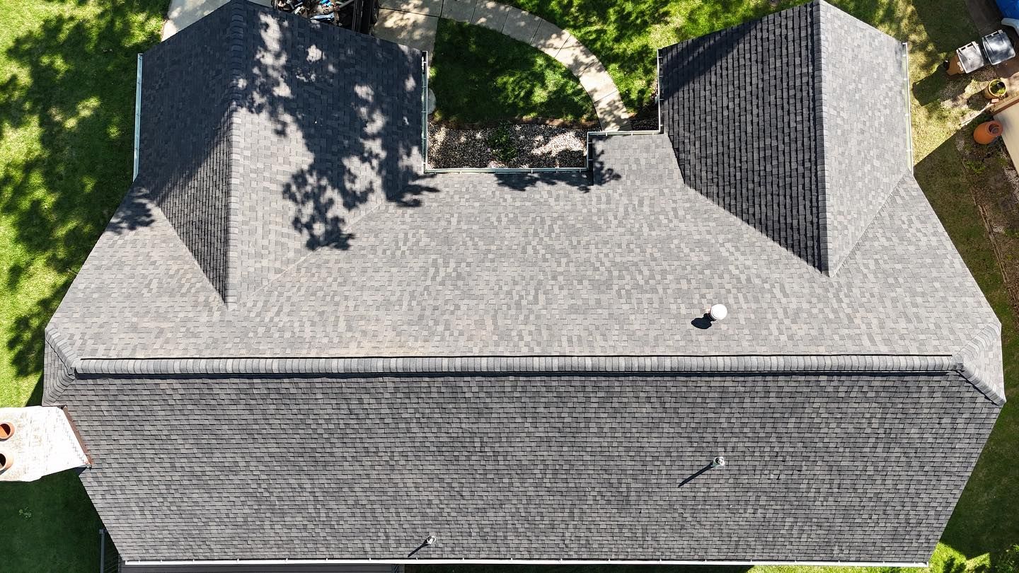 Overhead view of a house roof with dark gray shingles, dormers, and a shadow from a tree.