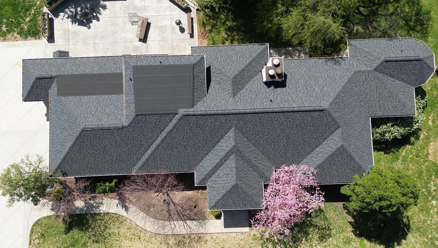 Aerial view of a dark gray shingle roof on a house, with a chimney and surrounding trees.