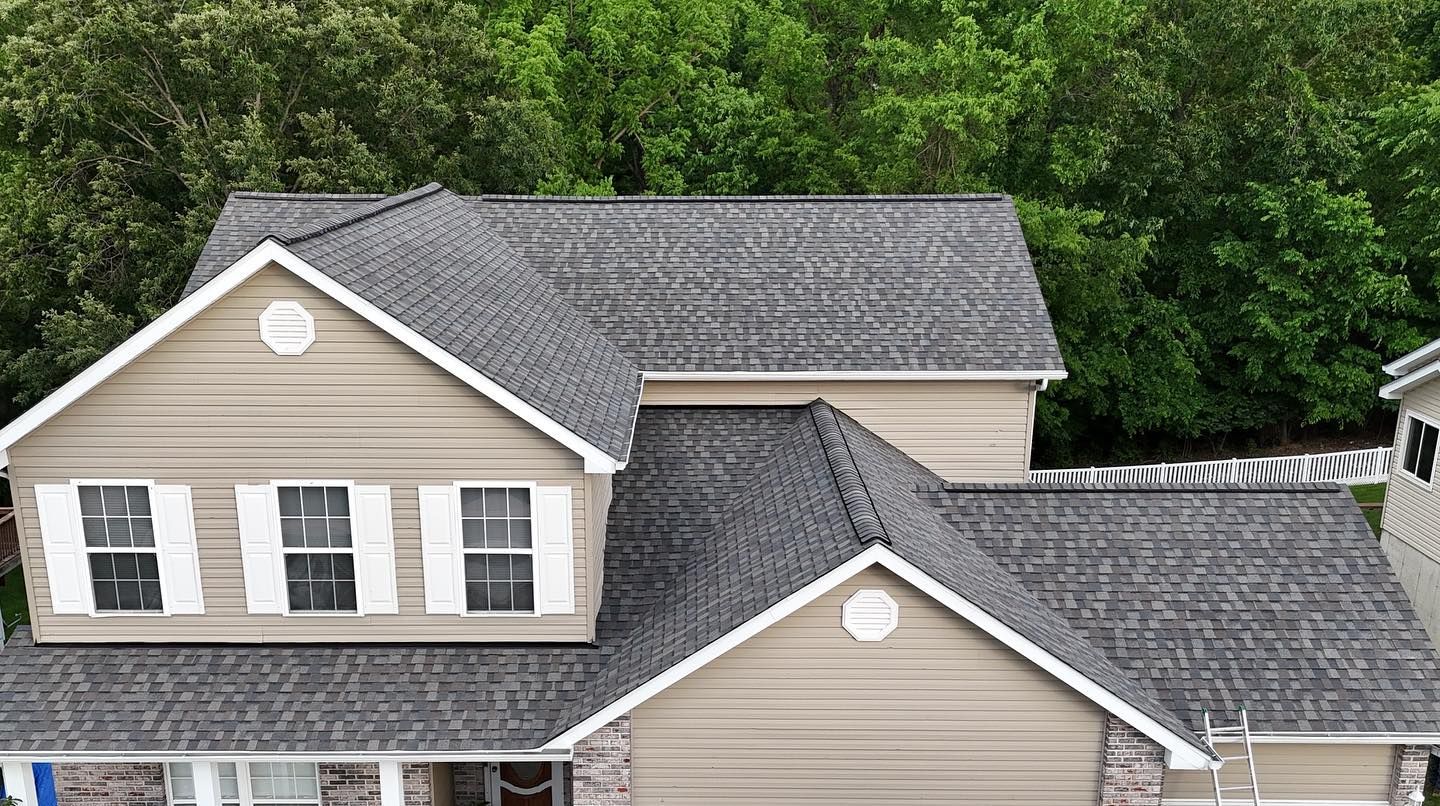 Tan house with dark gray roof, two windows, and lush green trees in background.