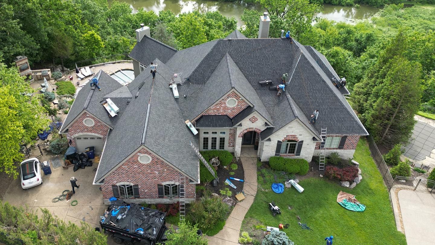 House with roofing crew, gray roof, brick and white siding, green yard, river in background.