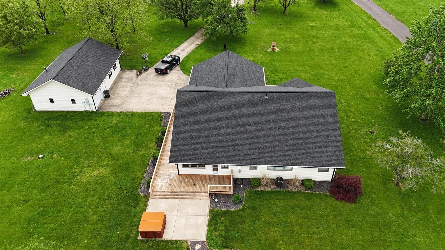 Aerial view of a white house with a dark gray roof, adjacent to a garage and surrounded by green grass and trees.