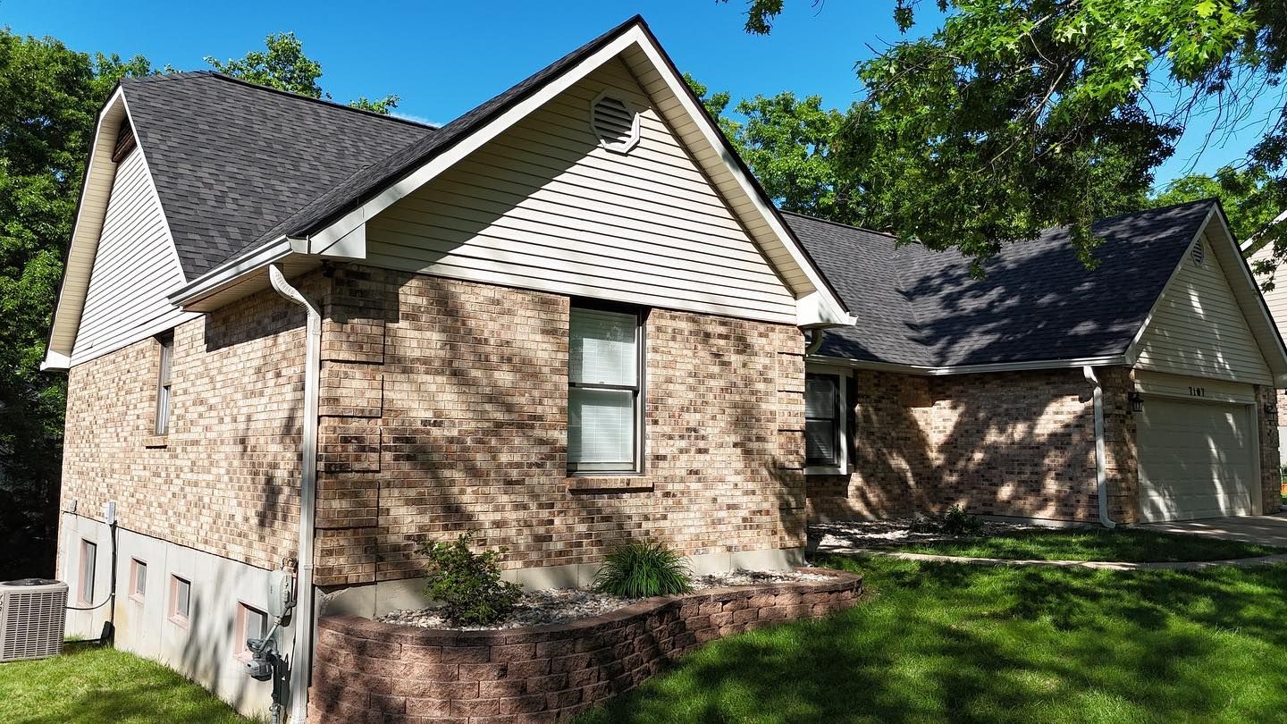 Brick house with dark roof and white trim, surrounded by green grass and trees on a sunny day.