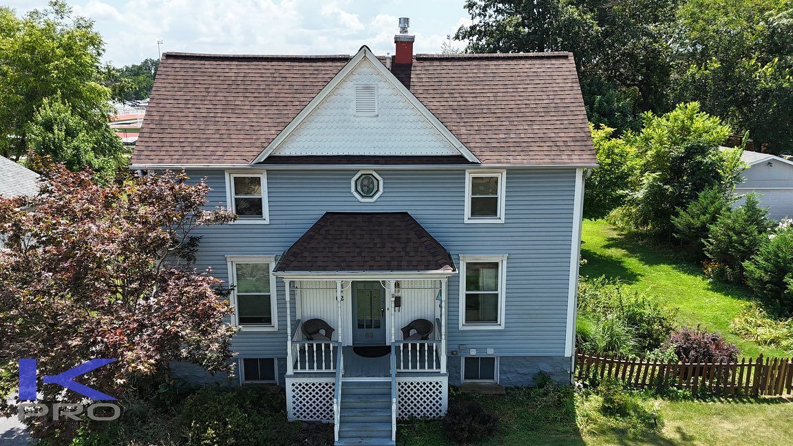 Blue two-story house with a porch and brown roof, set against a backdrop of trees and a sunny sky.