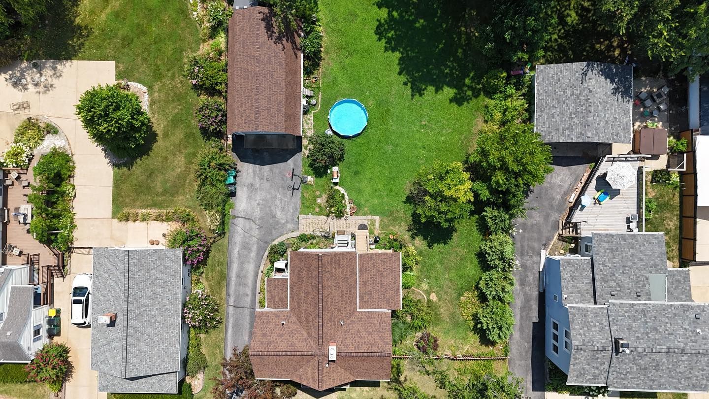 Aerial view of houses with brown roofs, driveways, and green lawns, with a blue swimming pool.