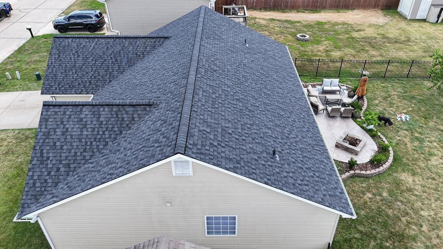 Overhead view of a house with a dark gray shingled roof and tan siding, surrounded by green grass and a small patio.