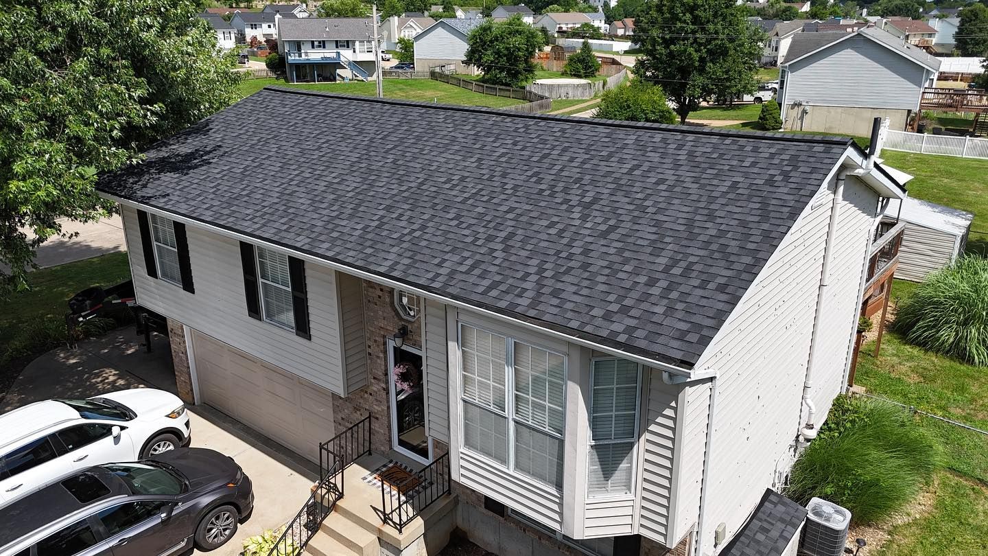 Two-story house with dark gray roof, two cars parked in front, surrounded by green grass and trees.