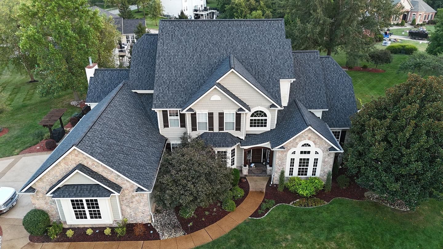 A large, two-story house with a gray roof and stone facade, surrounded by green grass and trees.