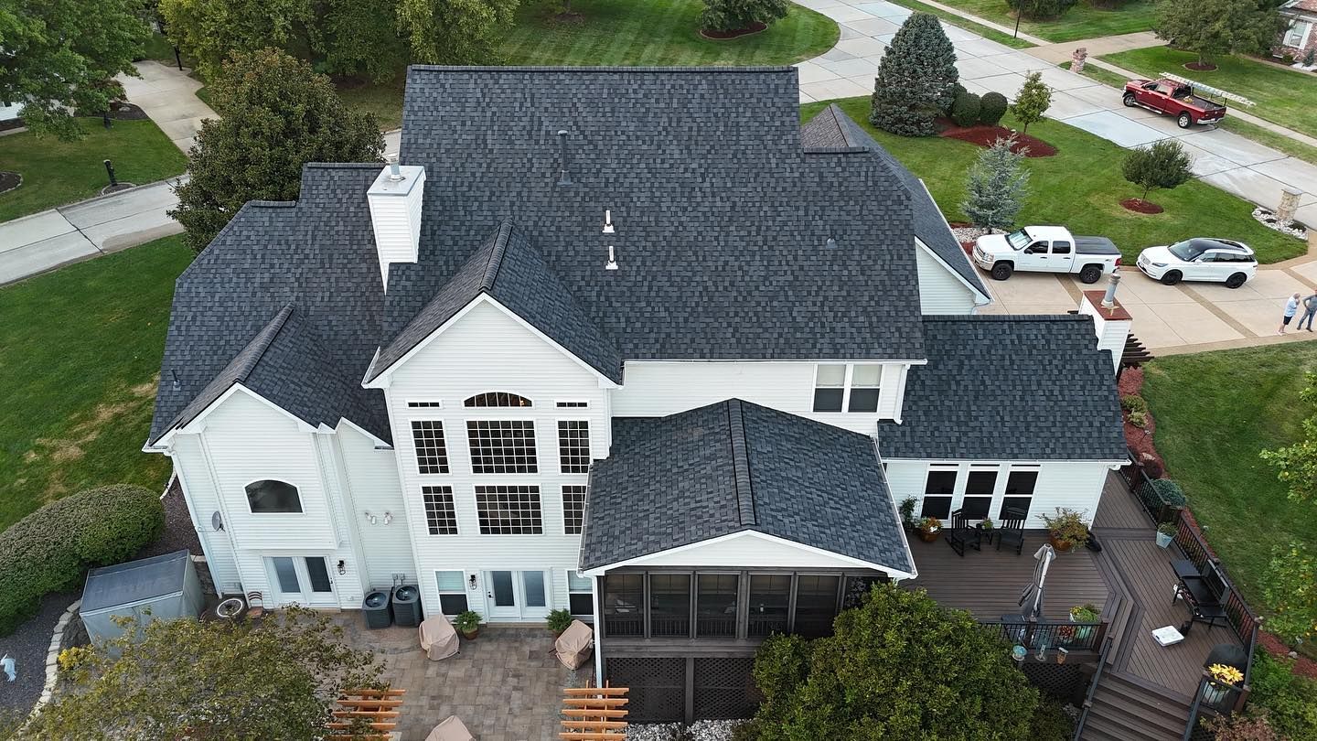 Aerial view of a white house with a dark gray roof, lush green lawn, and vehicles parked on the side.