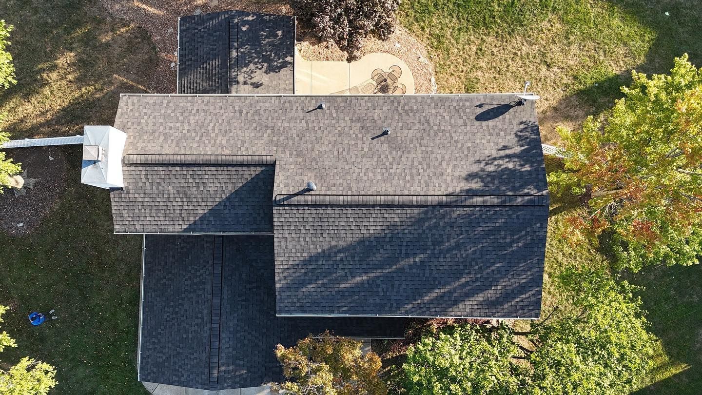 Overhead view of a dark gray shingled roof on a house, surrounded by trees and grass.
