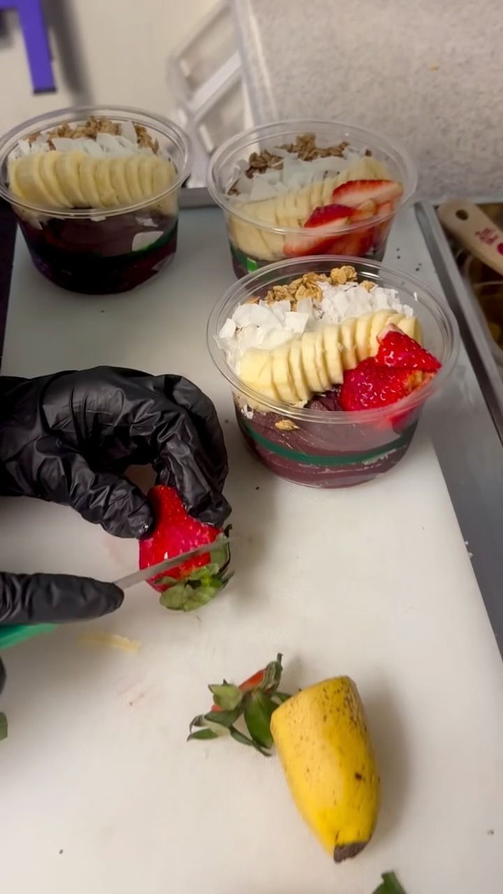 A person in black gloves slicing a strawberry, preparing a layered fruit dessert in plastic cups.