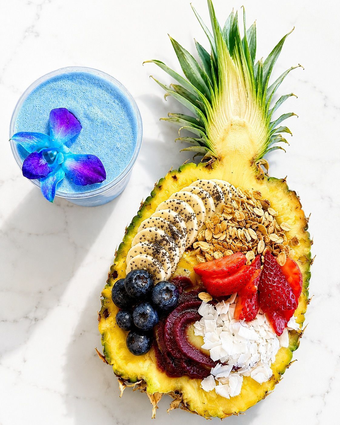 Pineapple bowl with yogurt, fruit, granola, and coconut; blue smoothie beside it on a white surface.