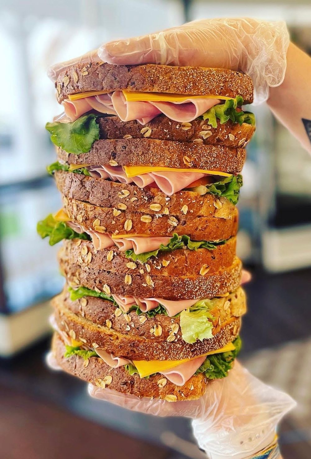 Gloved hand holding a tall stacked sandwich with lettuce, cheese, and meat layers
