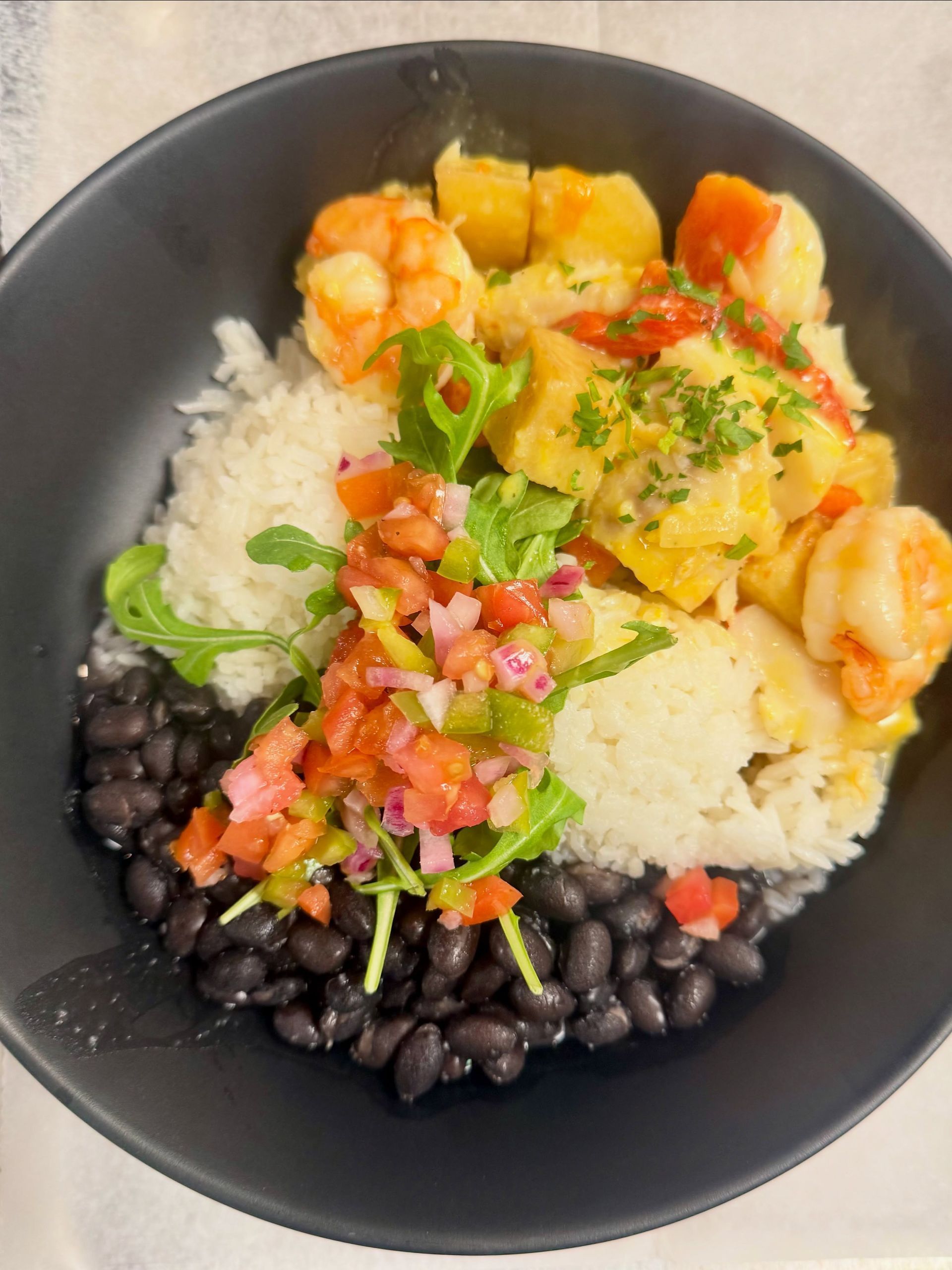 Black bowl with rice, black beans, shrimp, and colorful vegetables topped with salsa and herbs