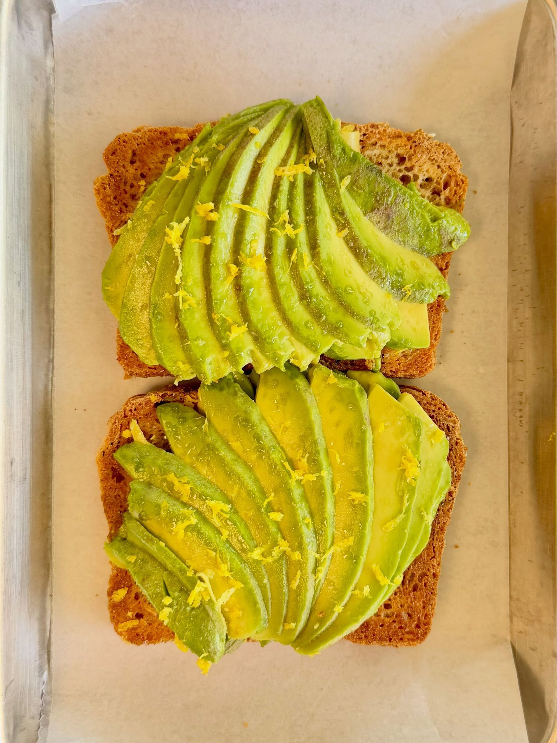 Two avocado toast slices in a white box, topped with fanned avocado on brown bread