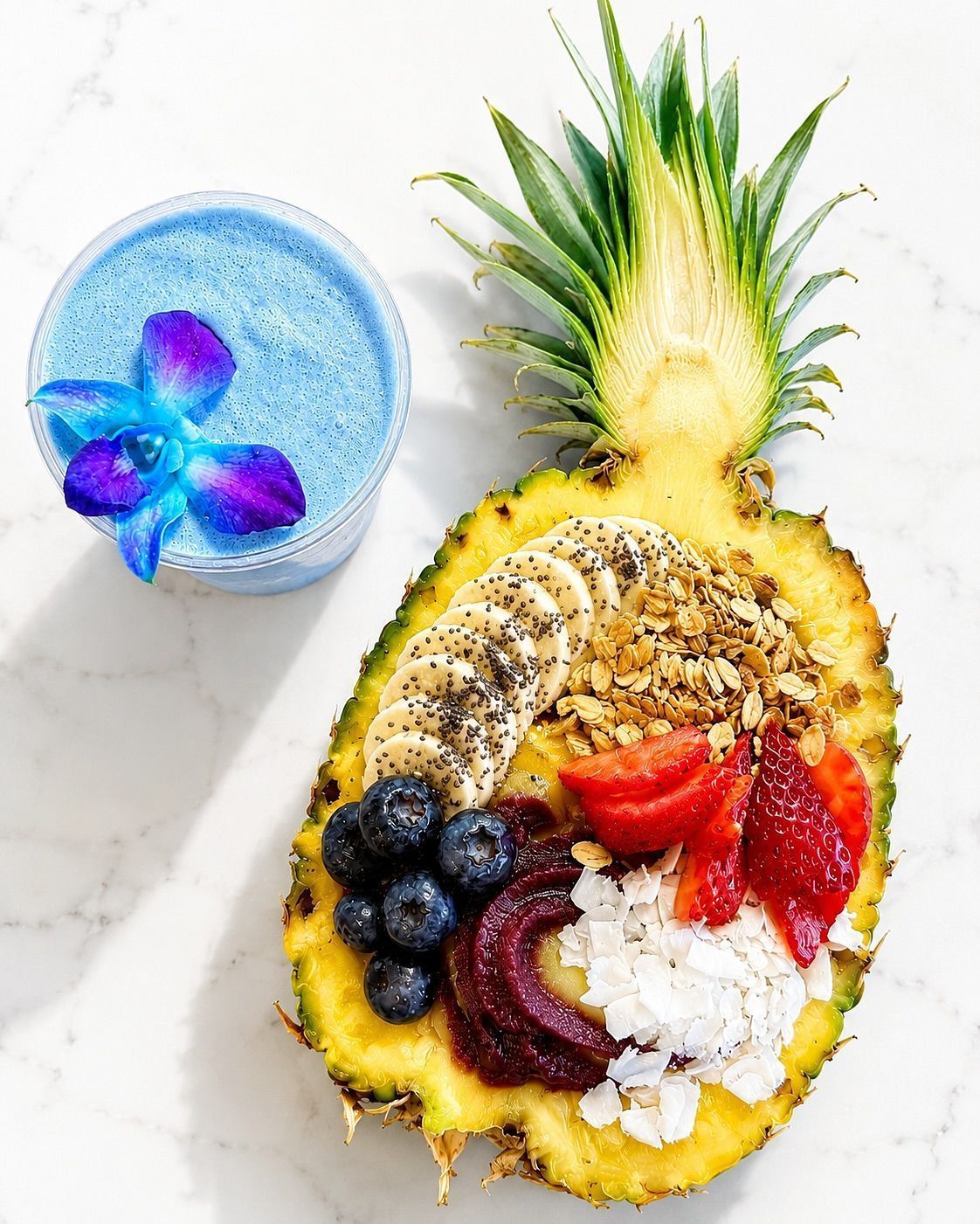 Pineapple bowl of fruit and granola beside a blue butterfly pea drink on a white surface
