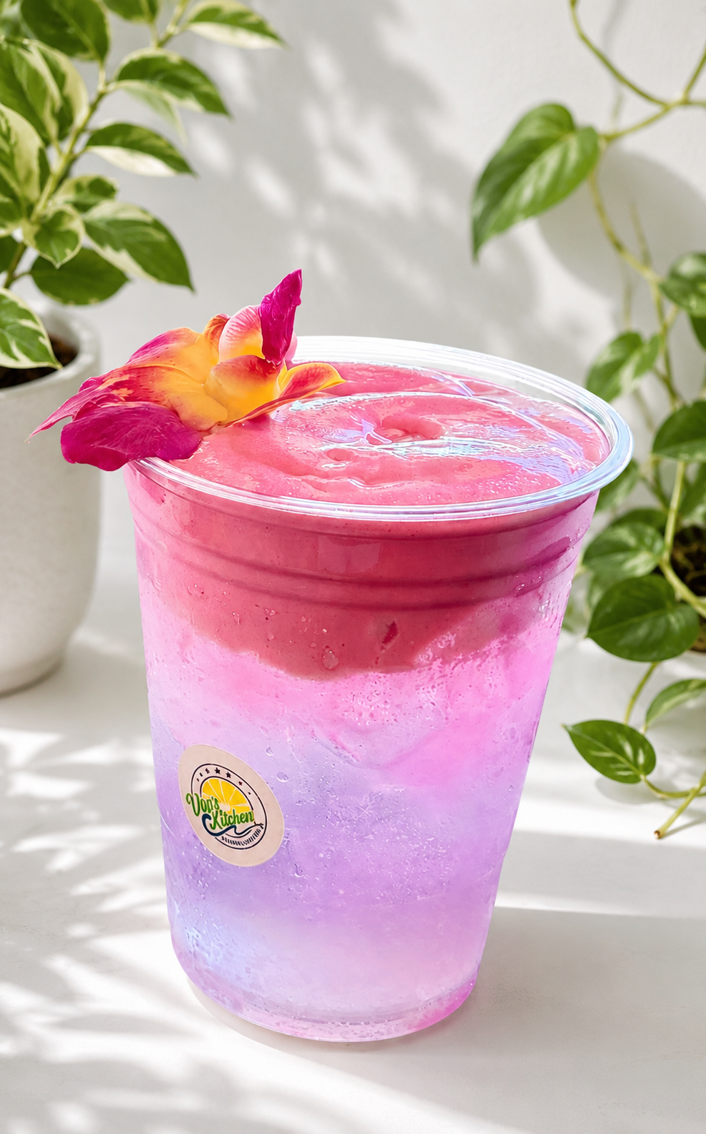 Pink-purple layered drink in a clear plastic cup, garnished with a flower, on a bright table with plants.