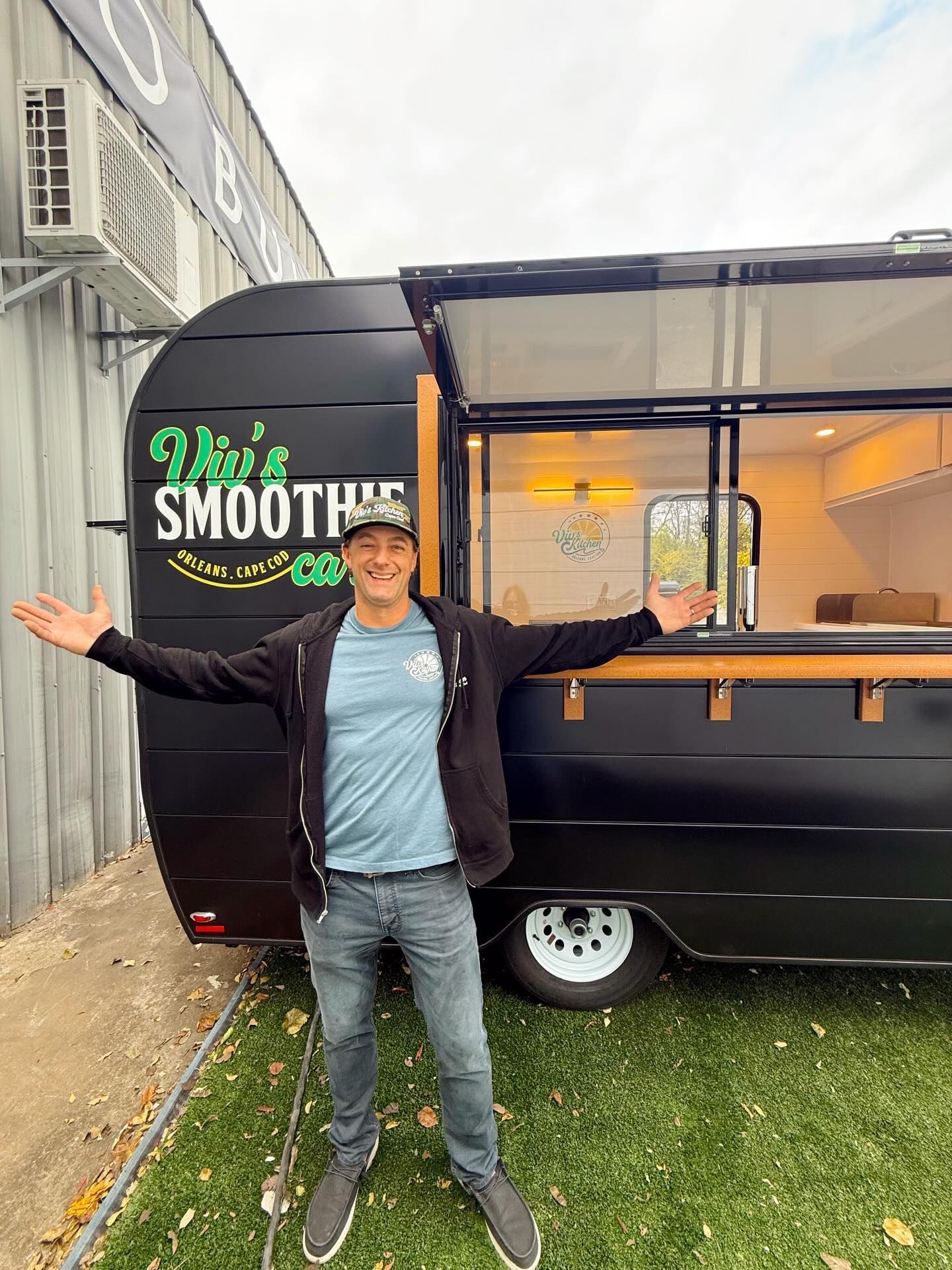 Person standing beside a black smoothie trailer with arms open and a Smoothie sign.